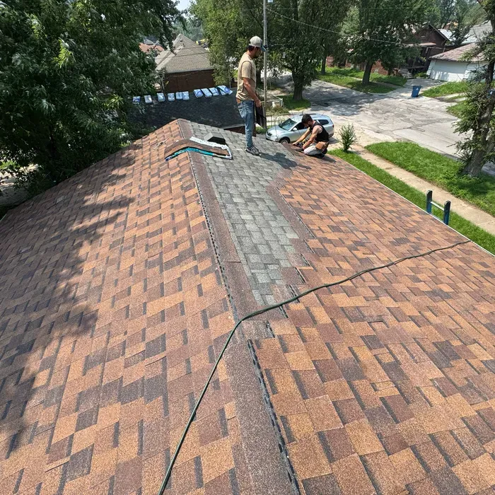 Workers on a brown shingle roof repair a section near a chimney.