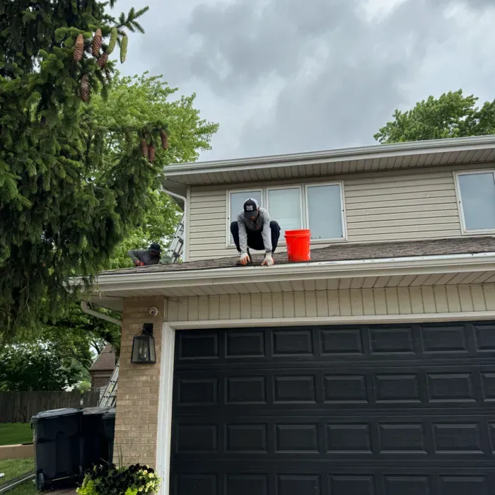 Person crouching on roof near a red bucket, working on a house with a black garage door.