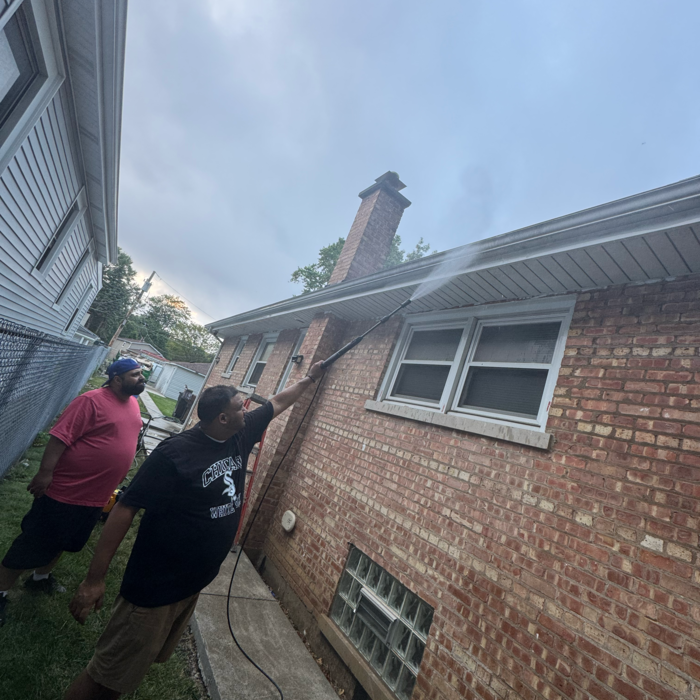 Two people power washing a brick house; one sprays the siding. Cloudy sky, backyard setting.