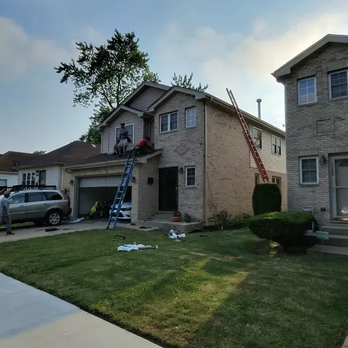 Workers on ladders at a brick house, likely roof repair. Green lawn, blue sky, parked car, and adjacent house visible.