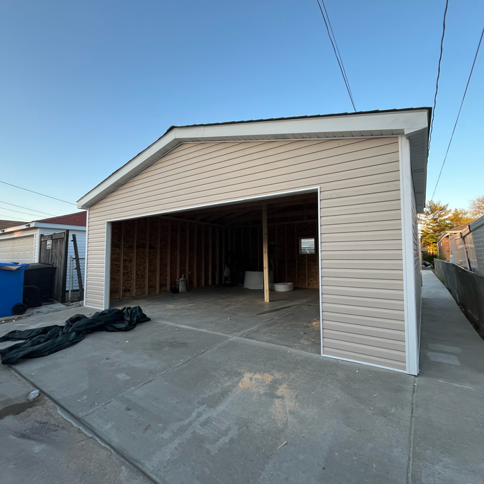Newly constructed beige vinyl-sided garage with open entrance. Concrete driveway, blue sky.