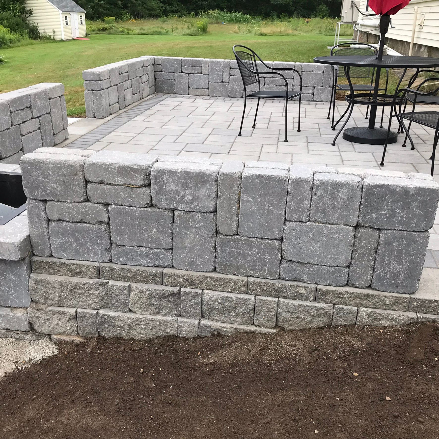 Stone patio with built-in seating area, gray pavers, black furniture, and surrounding greenery.