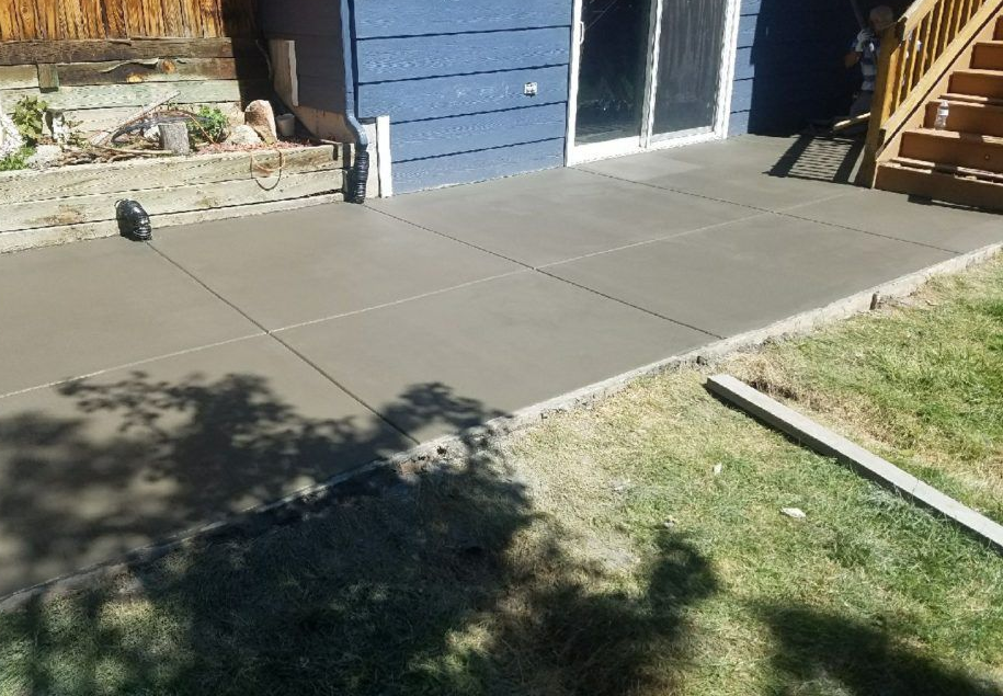 Newly poured concrete patio next to blue house with sliding door and stairs, next to a grass yard.