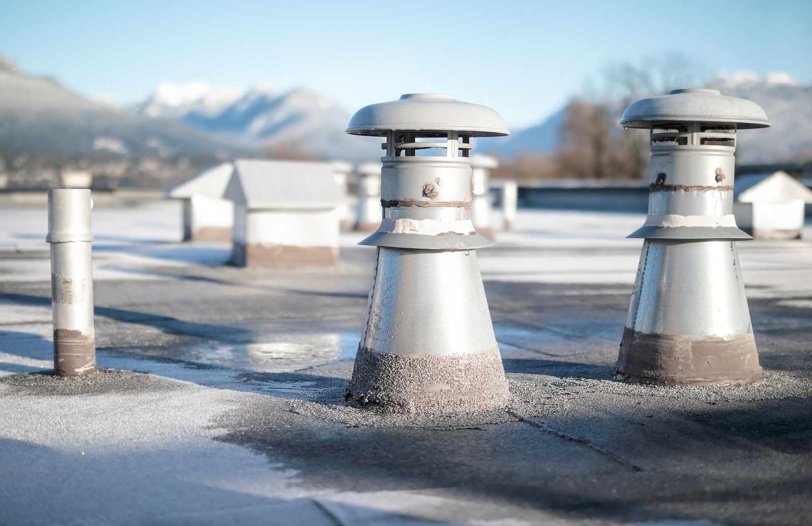 Rooftop with three metal vents and small buildings, with snowy mountains in the background.