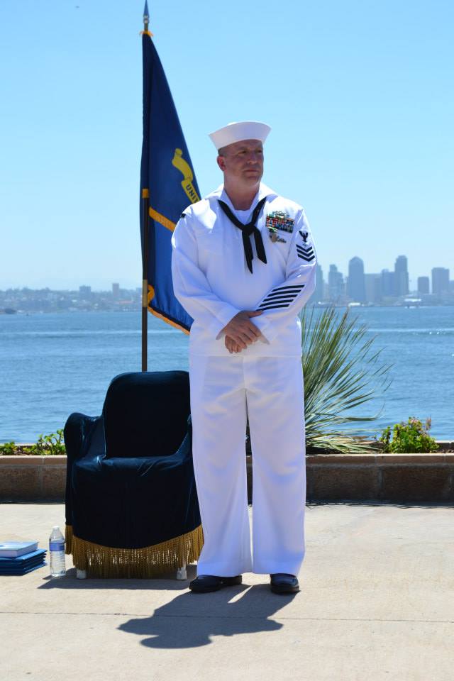 A man in a sailor uniform stands in front of a flag