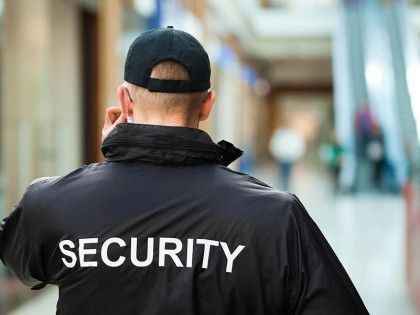 A security guard is talking on a cell phone while wearing a black jacket and hat.