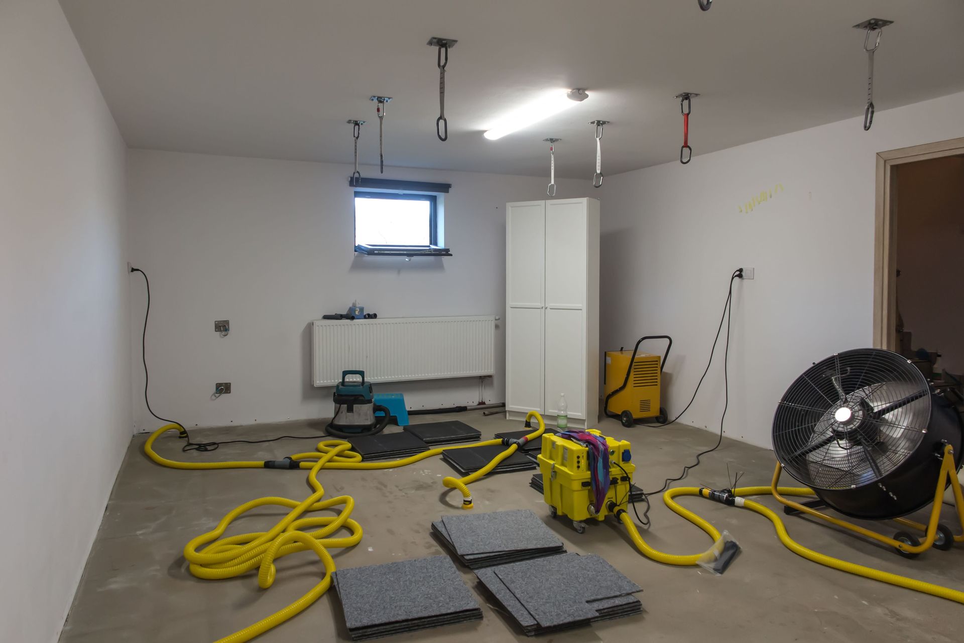 A room with drying equipment: fans, hoses, and dehumidifiers. White walls and grey flooring.