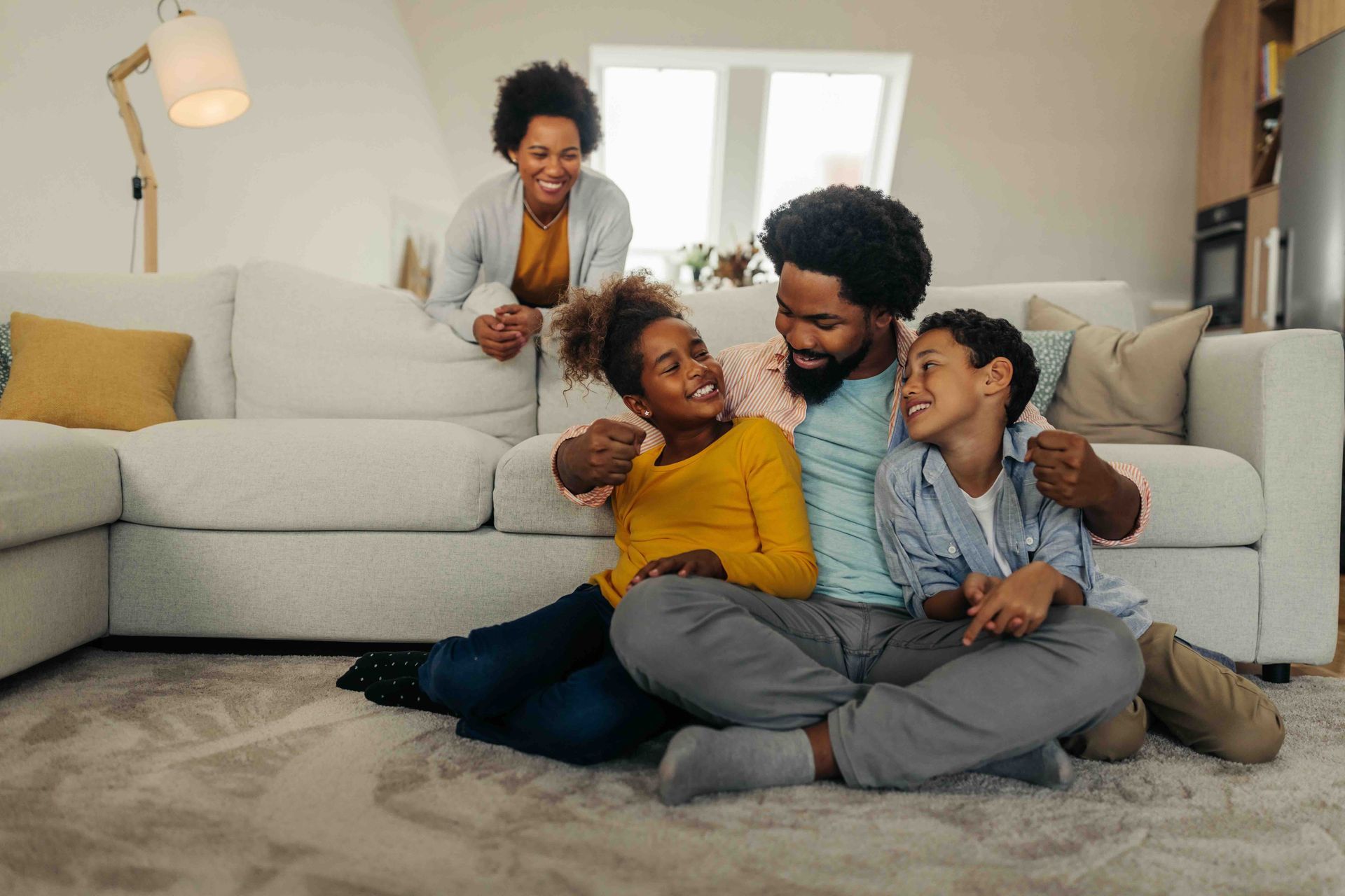 Family smiling together on a living room floor; woman leans on the couch, two children and a man.