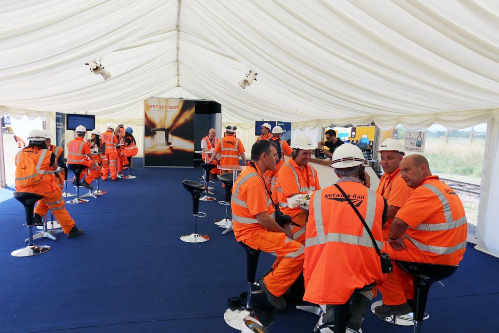 A group of construction workers are sitting at tables under a tent.