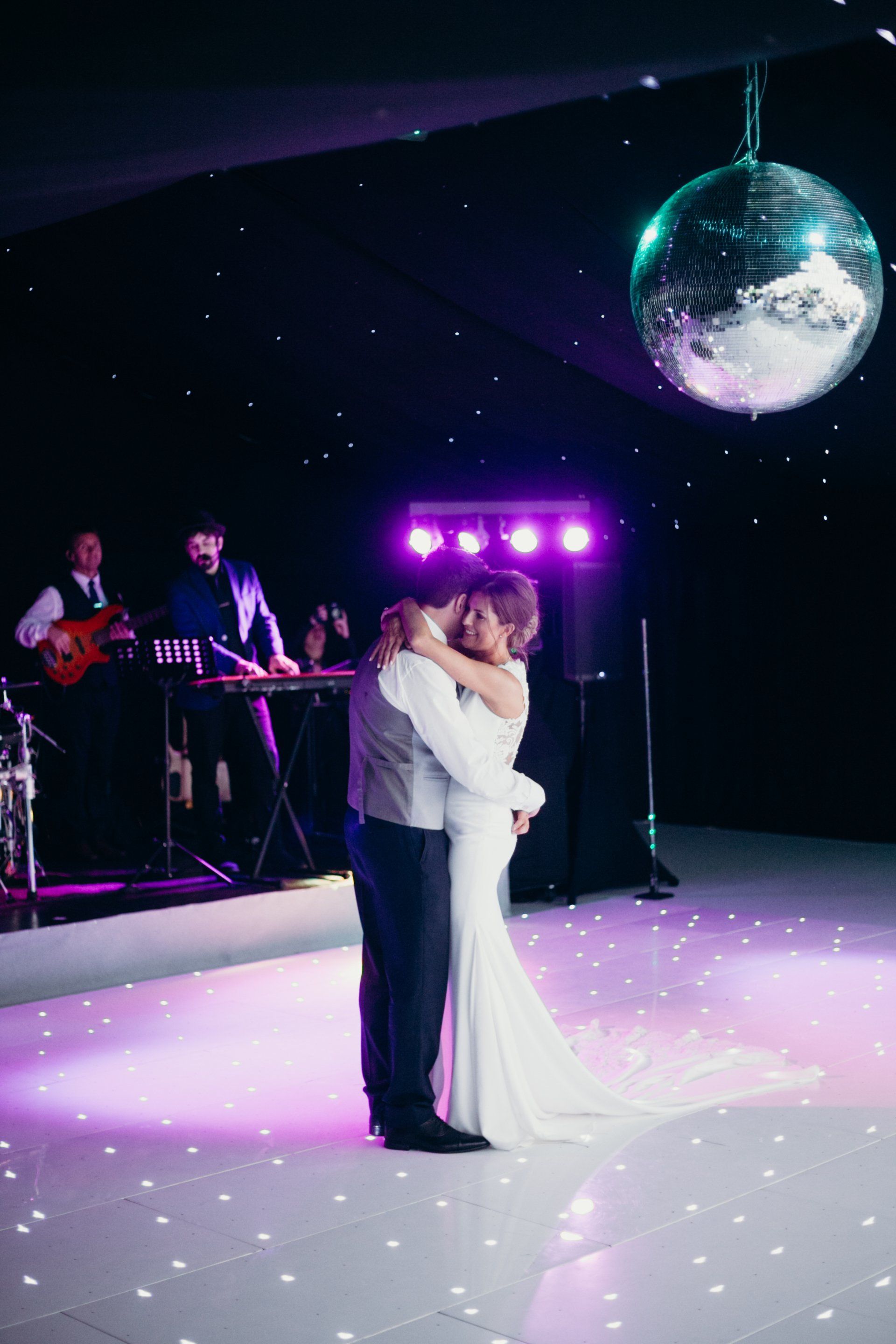 A bride and groom are dancing on a white dance floor with a disco ball in the background.