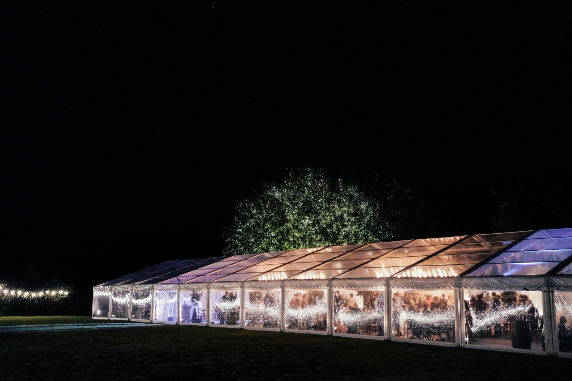 A clear tent is lit up at night with a tree in the background.