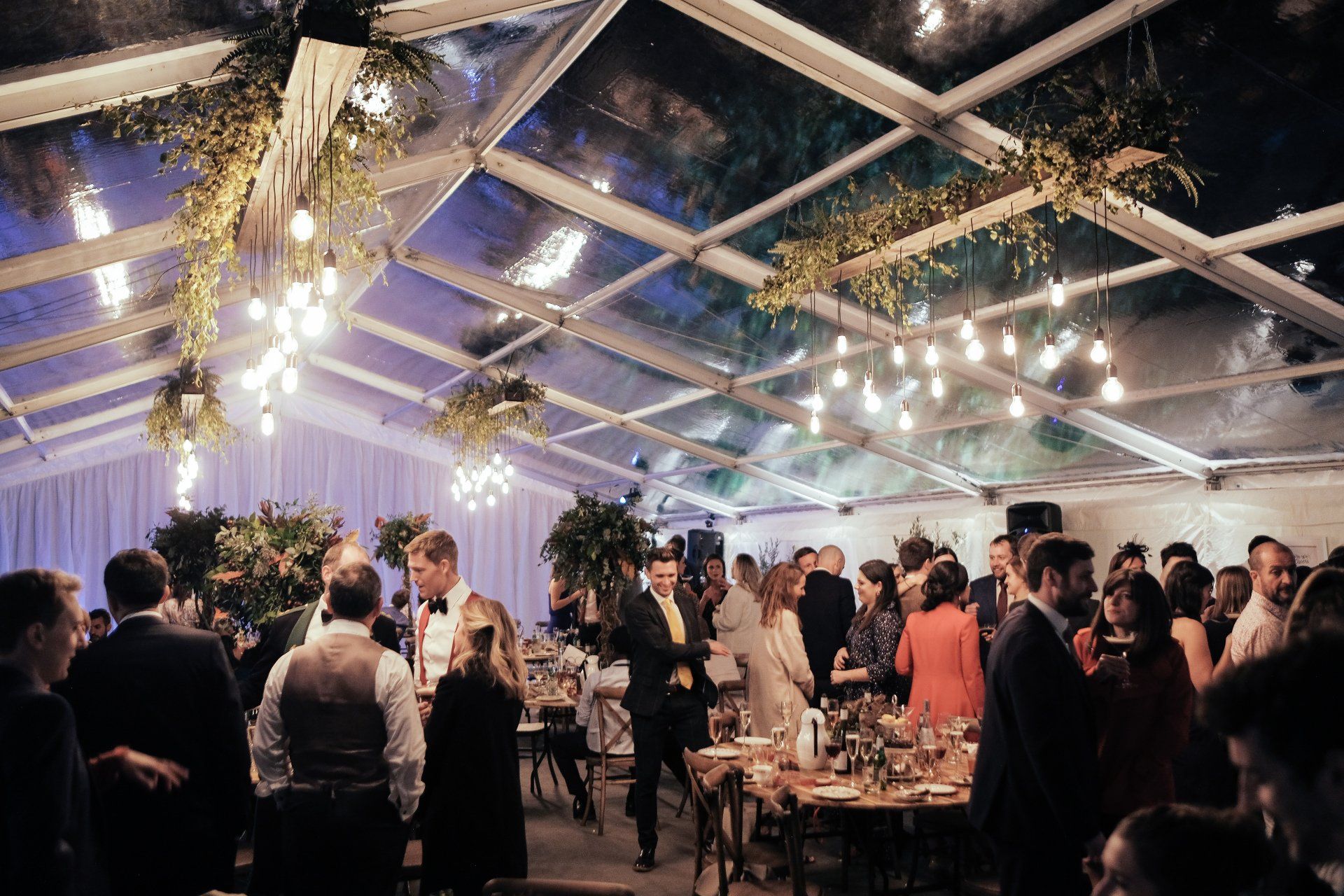 A large group of people are dancing under a clear tent at a wedding reception.