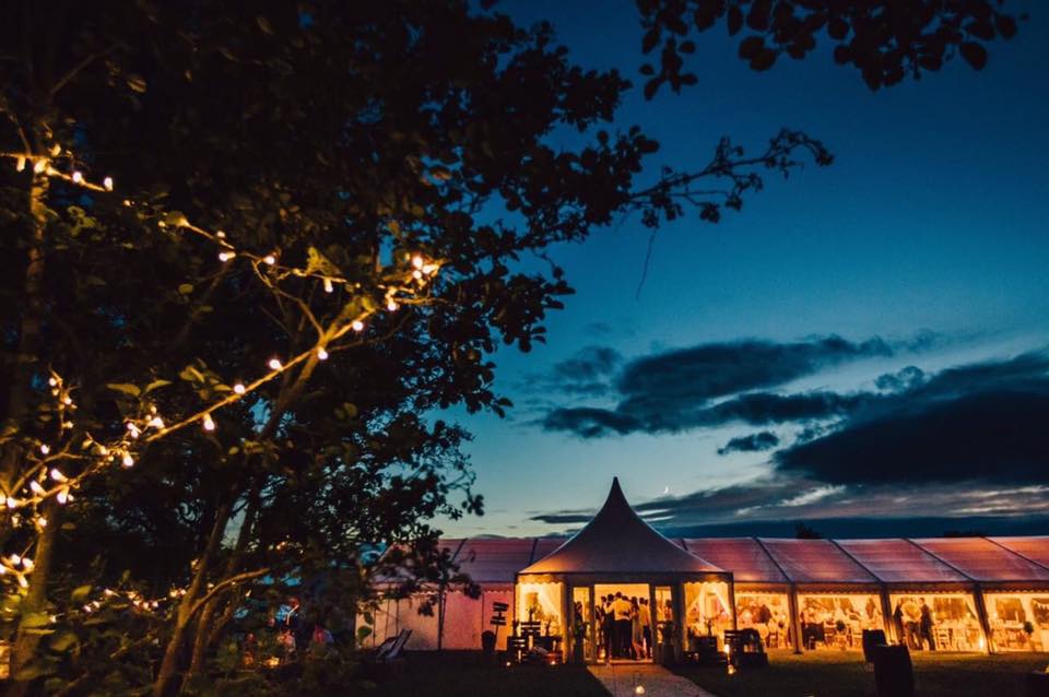 A large tent is lit up at night with trees in the foreground.