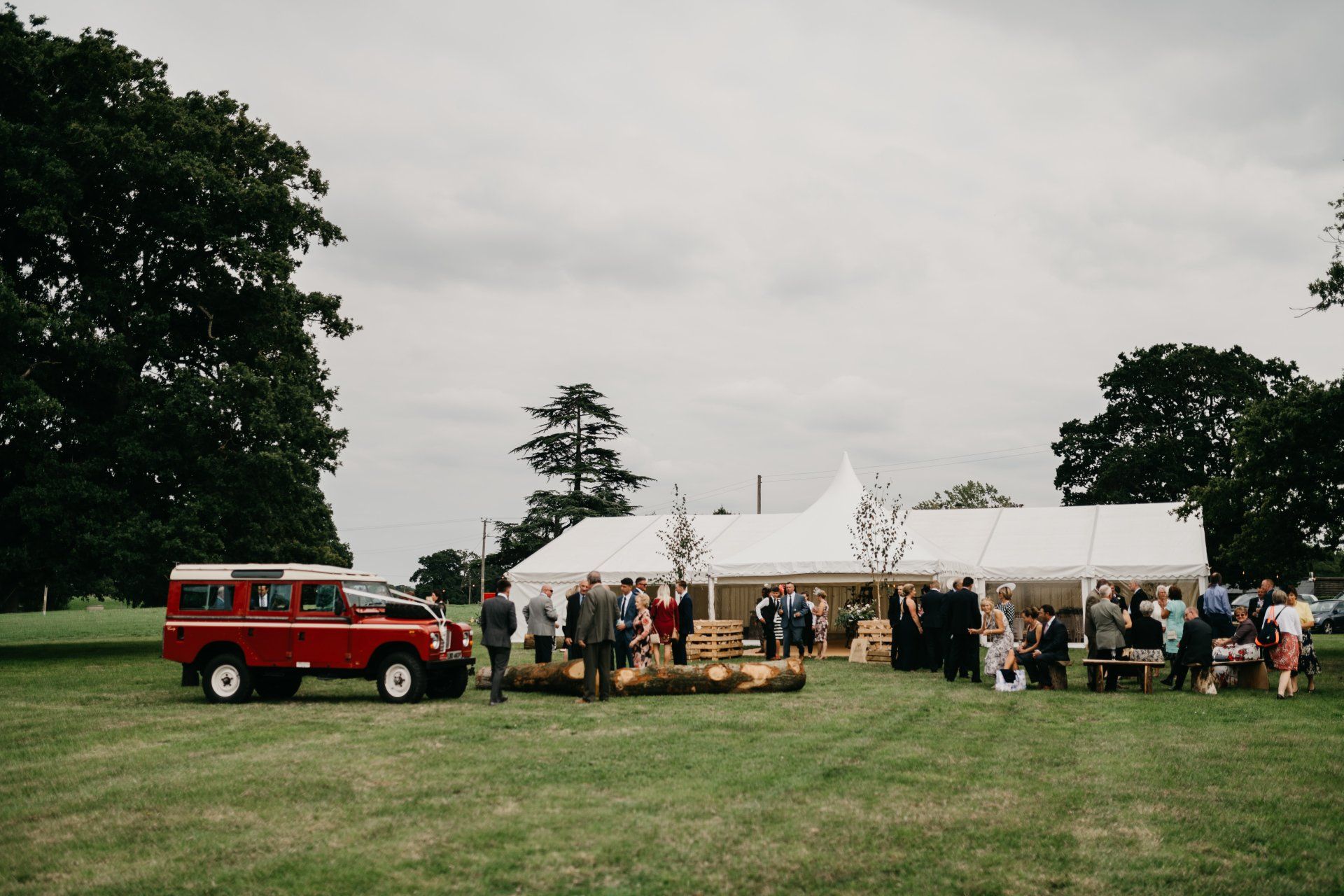 A red land rover is parked in a grassy field in front of a white tent.