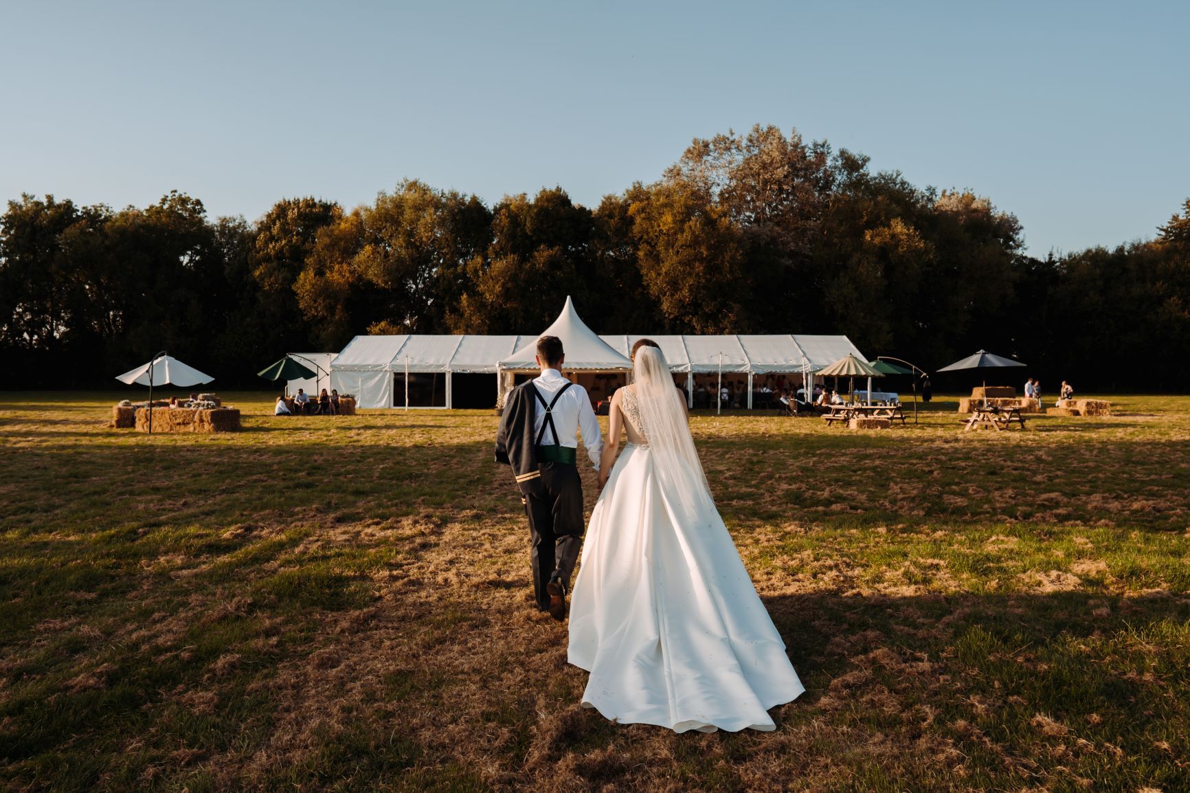 A bride and groom are walking in a field in front of a tent.