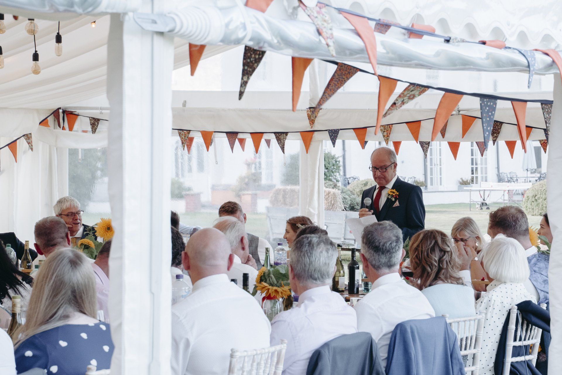 A man is giving a speech at a wedding reception in a tent.
