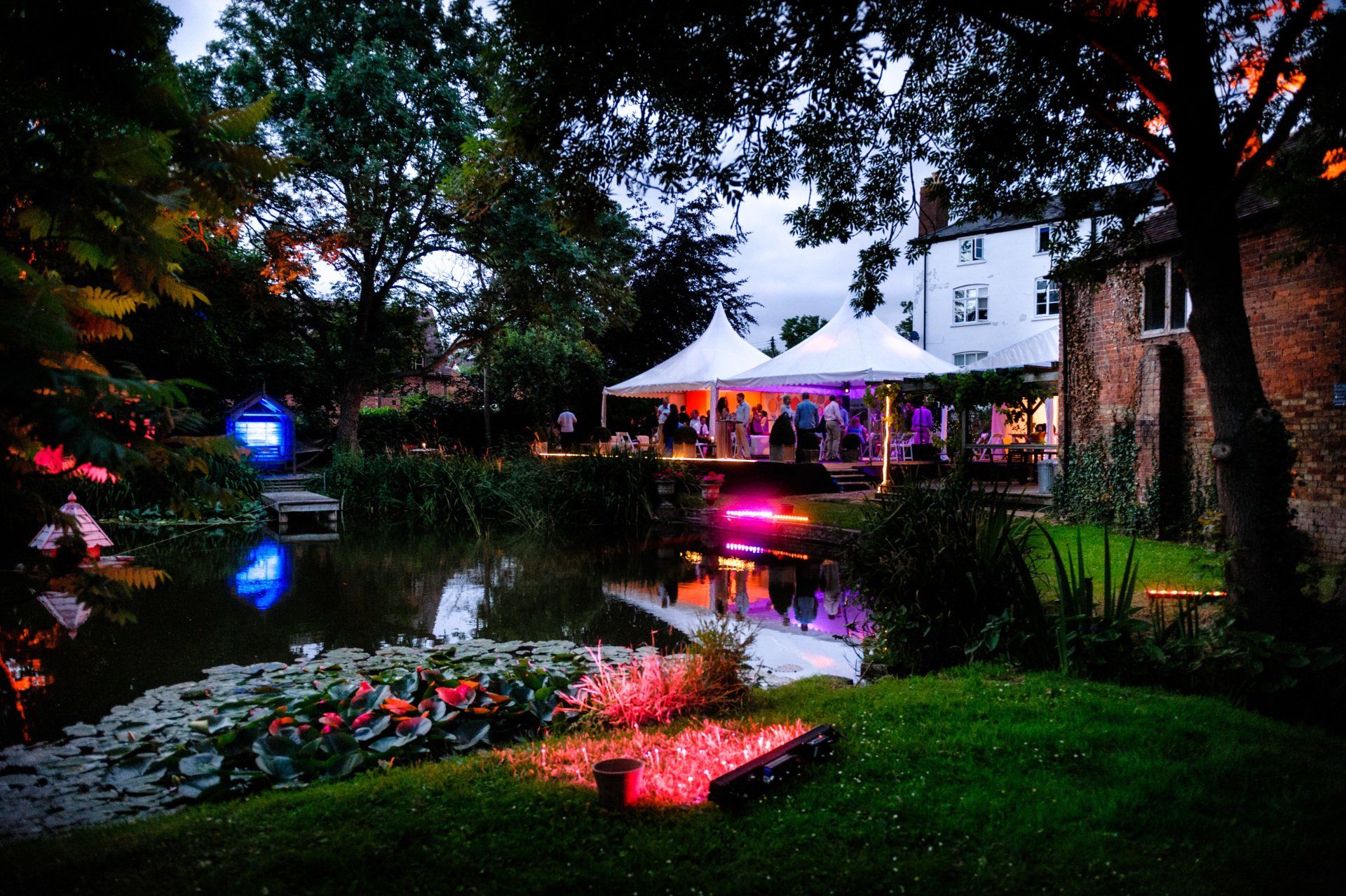 A tent is lit up in the middle of a garden next to a pond