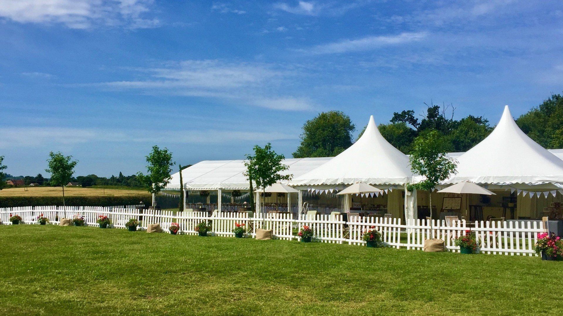 A row of white tents are sitting in a grassy field behind a white picket fence.