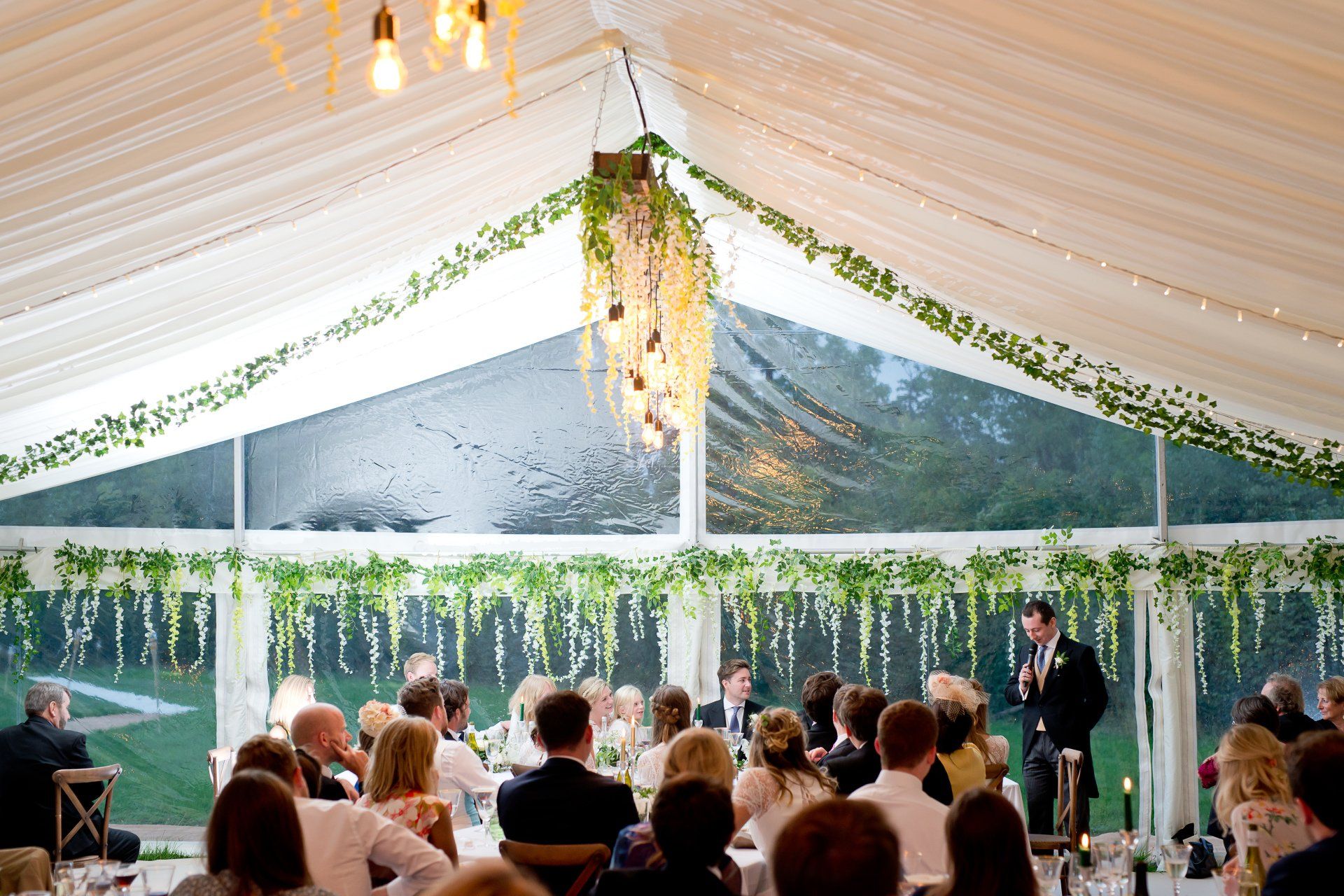 A man is giving a speech at a wedding reception in a tent.