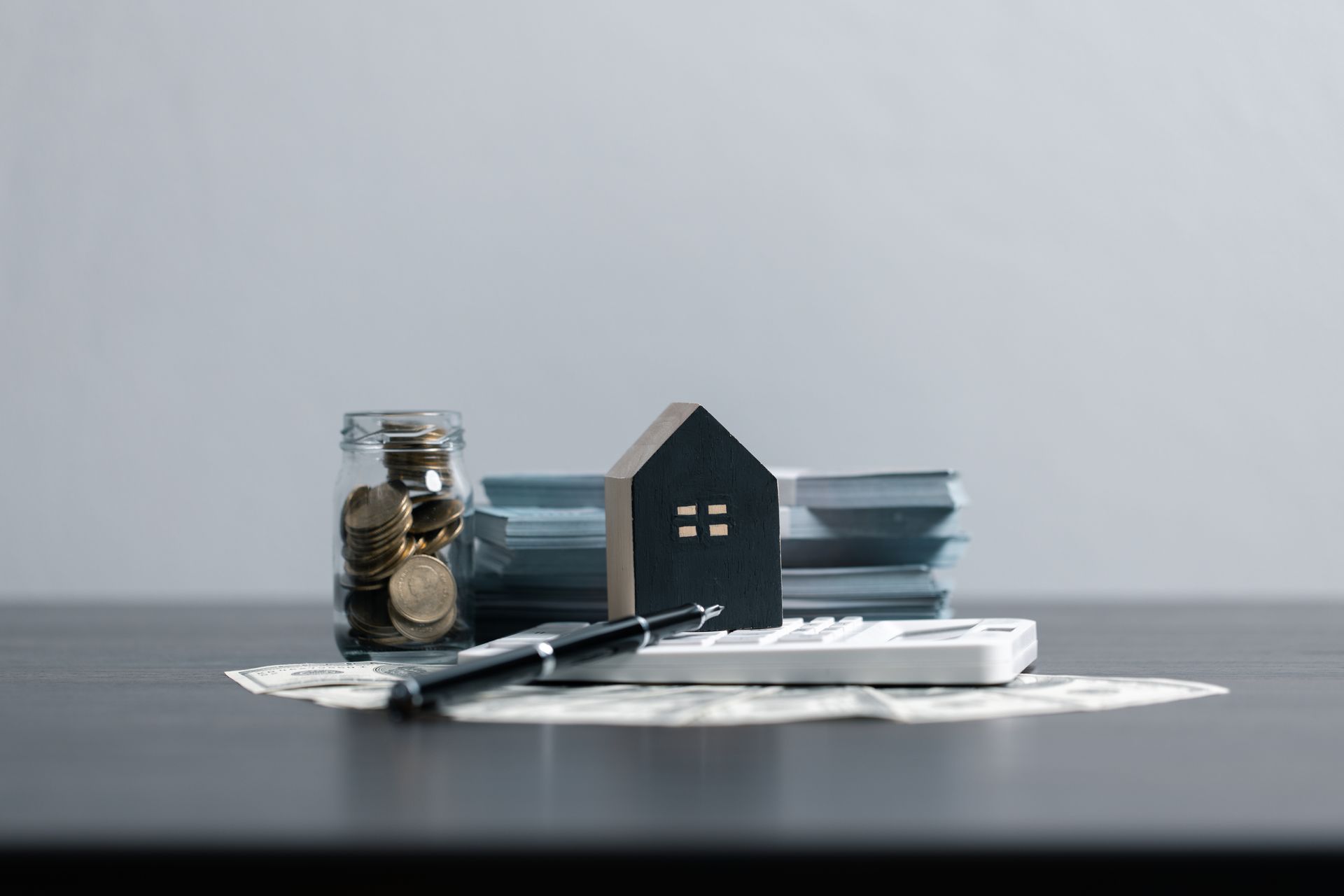 A small house , a calculator , a pen , and a jar of coins are on a table.