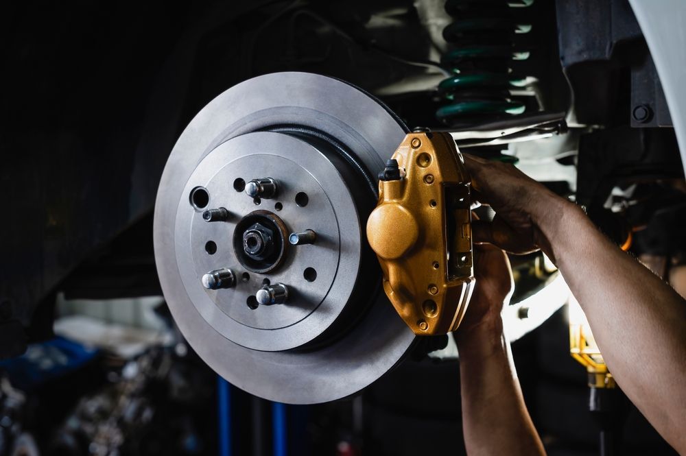 A mechanic works on a car's brake system, holding a yellow brake caliper. The brake rotor and suspension are also visible.