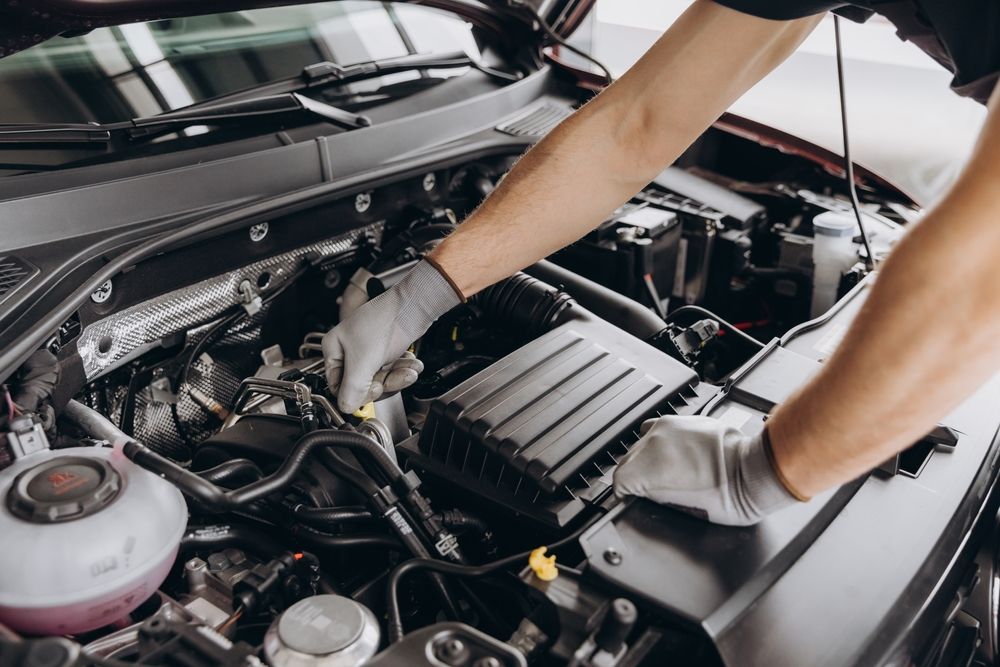 Mechanic wearing gloves working on a car engine, likely inspecting or repairing components in a garage.
