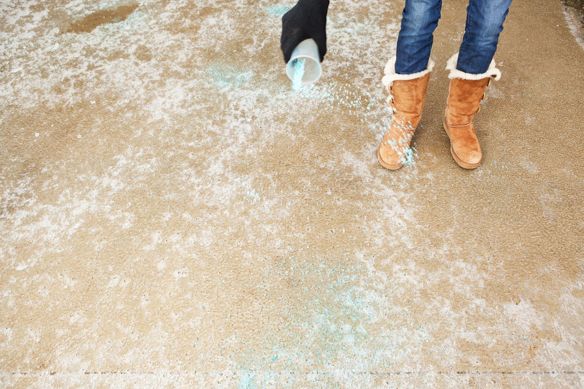Woman in brown boots sprays blue ice melt with a cup on a brown driveway.