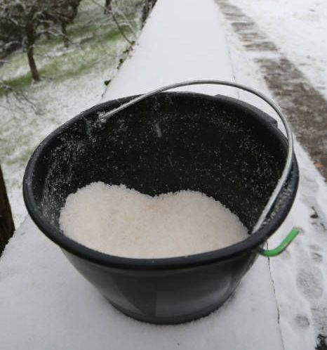 Black bucket filled with ice salt, with a blurry road with snow on the background.