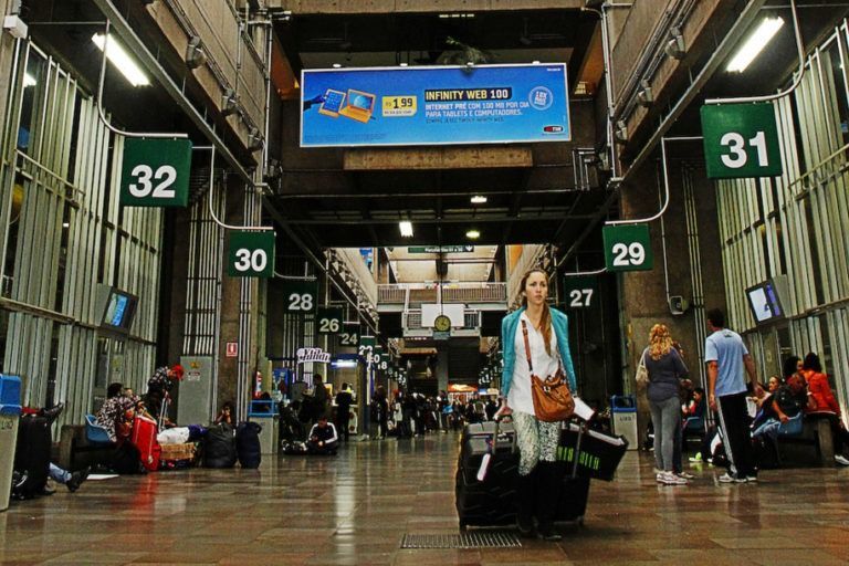 Mulher com bagagem caminhando por uma estação de trem movimentada, passando por plataformas numerada