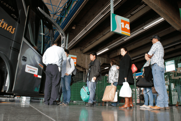 Pessoas embarcam em um ônibus em uma estação. Uma placa indica 