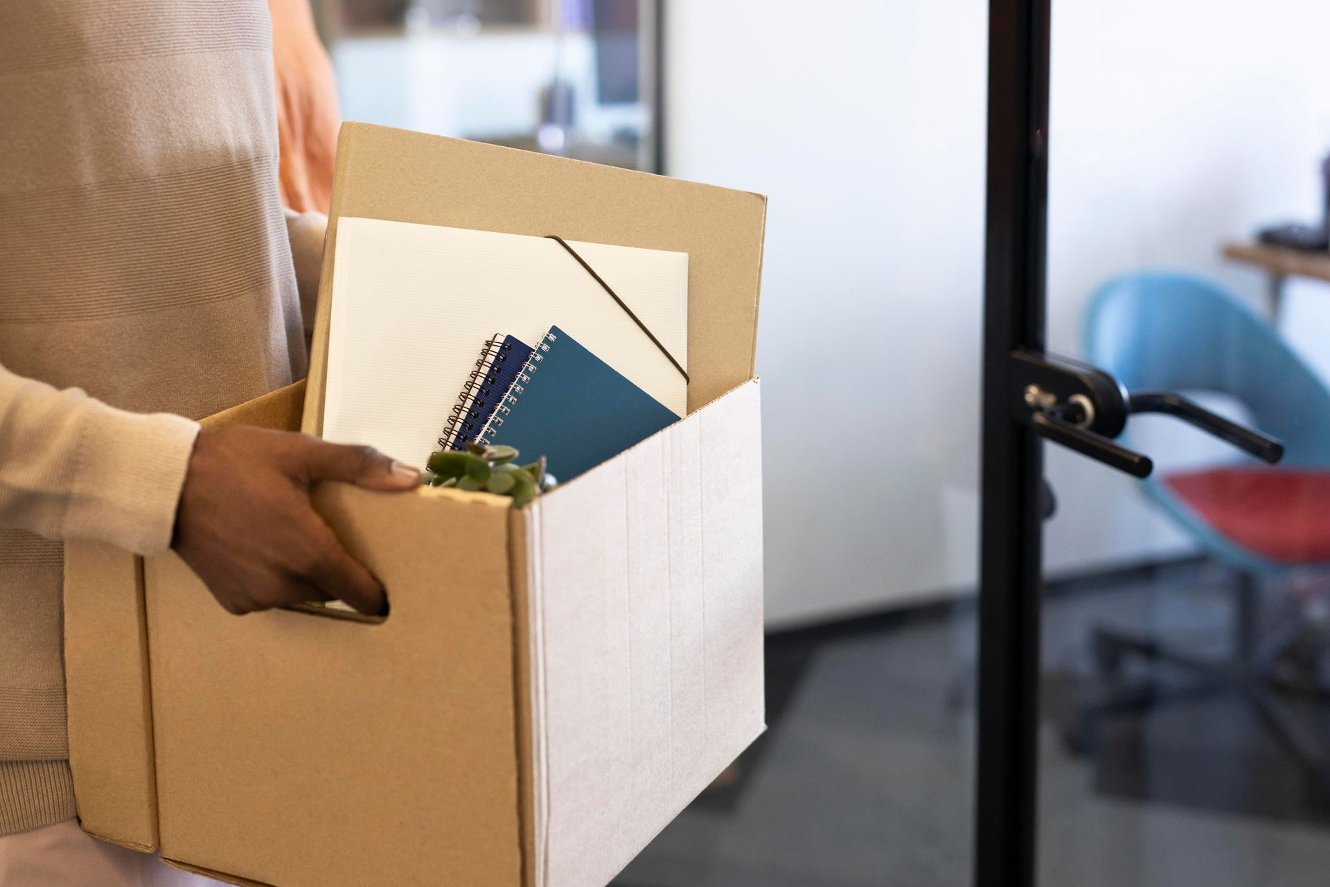 Pessoa carregando uma caixa de papelão com material de escritório, saindo do local de trabalho.