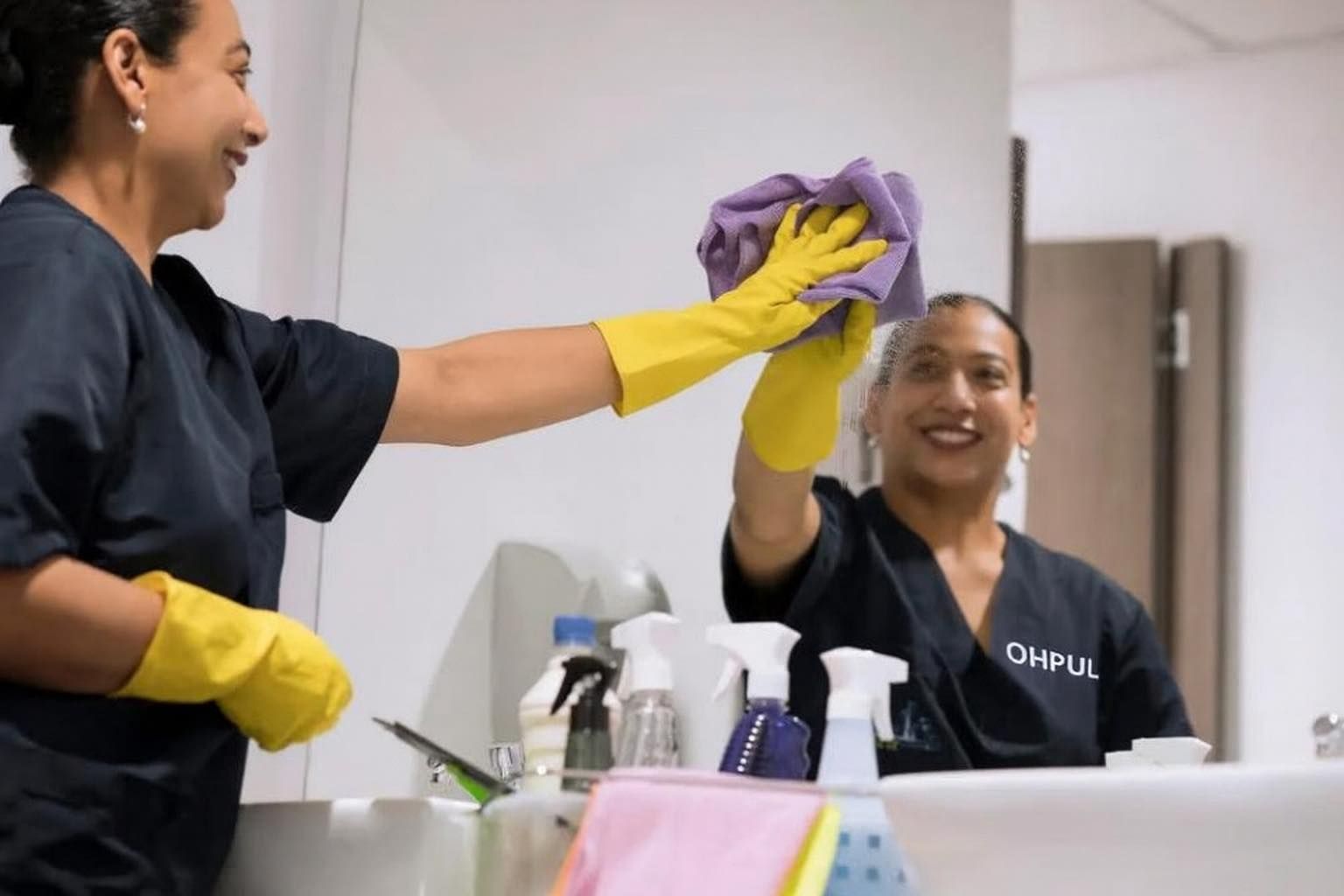 Two people cleaning a bathroom mirror. They wear gloves and smile.