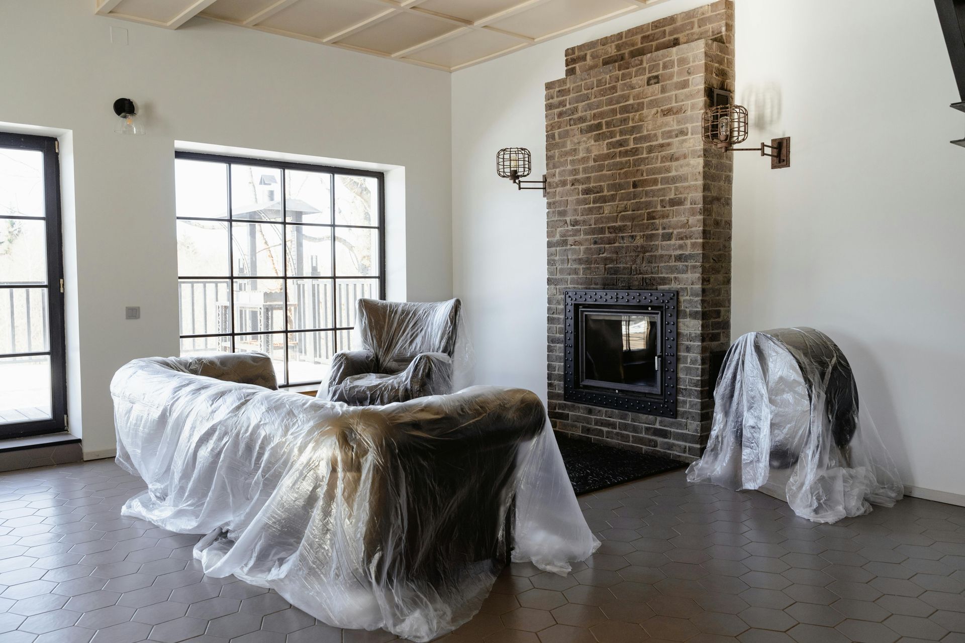 Living room with furniture covered in plastic, brick fireplace, large windows.