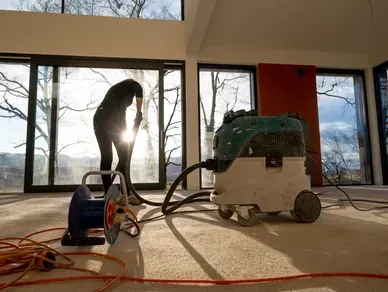 Person using a power tool, connected to a vacuum in a sunlit interior with large windows.