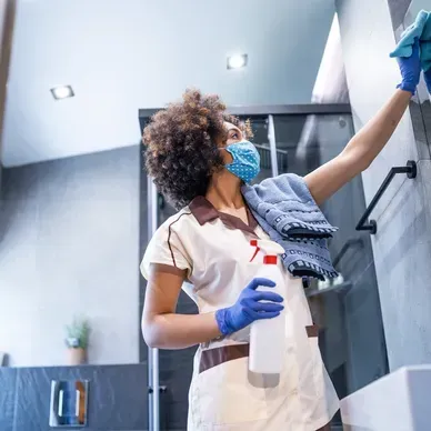 Person wearing a mask and gloves cleans bathroom wall with spray bottle and cloth.