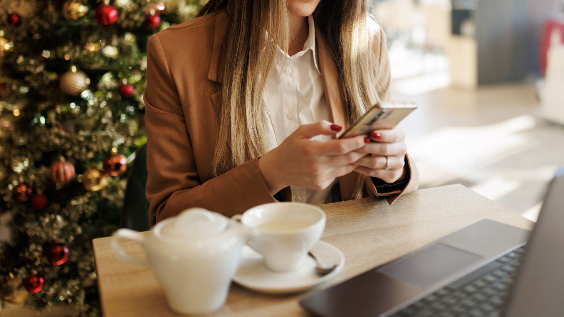 Woman using phone at table with tea and laptop, Christmas tree in background.