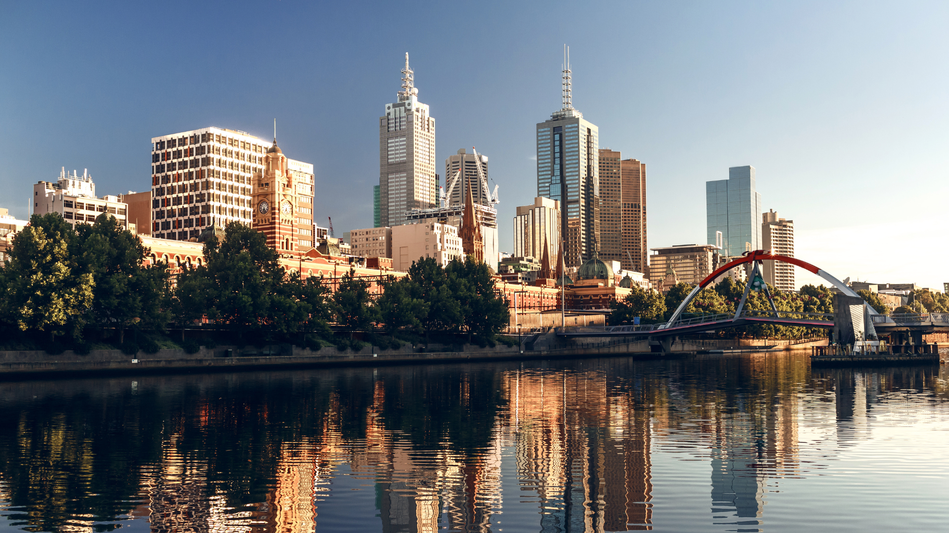 Melbourne skyline reflected in the Yarra River, featuring skyscrapers, trees, and a red bridge.