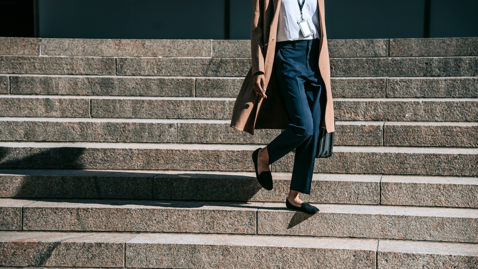 Woman descends stone steps outdoors wearing a tan coat, dark pants, and black flats.
