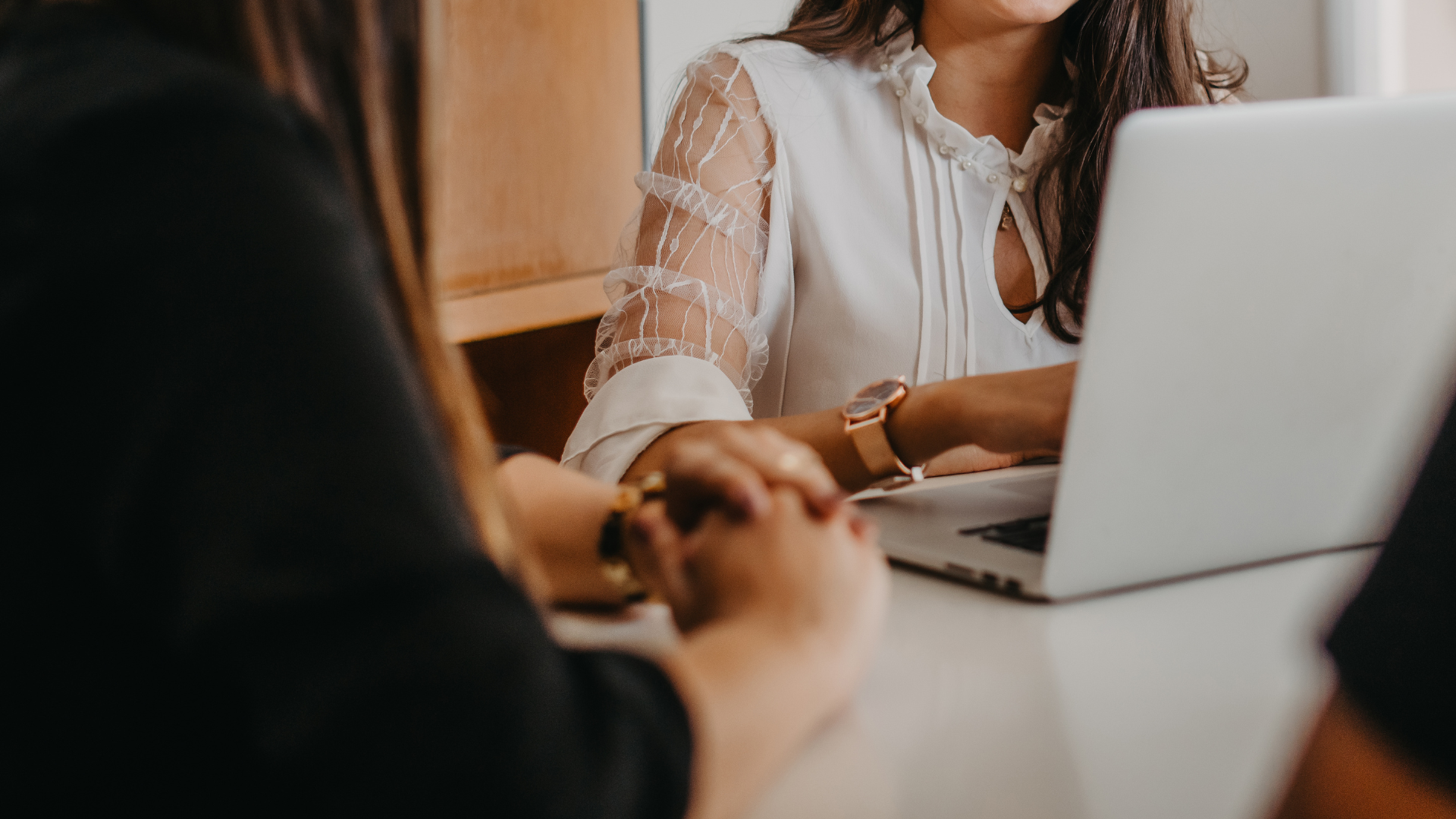 Three people at a table with a laptop, having a discussion. One woman wears a white blouse with intricate sleeves.