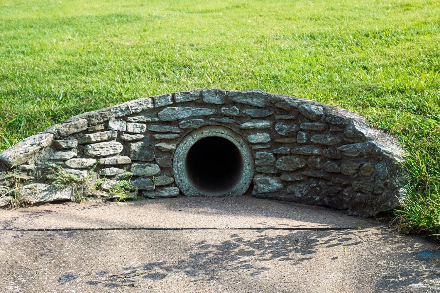 A round drainage pipe set into a stone retaining wall on the edge of a grassy field.