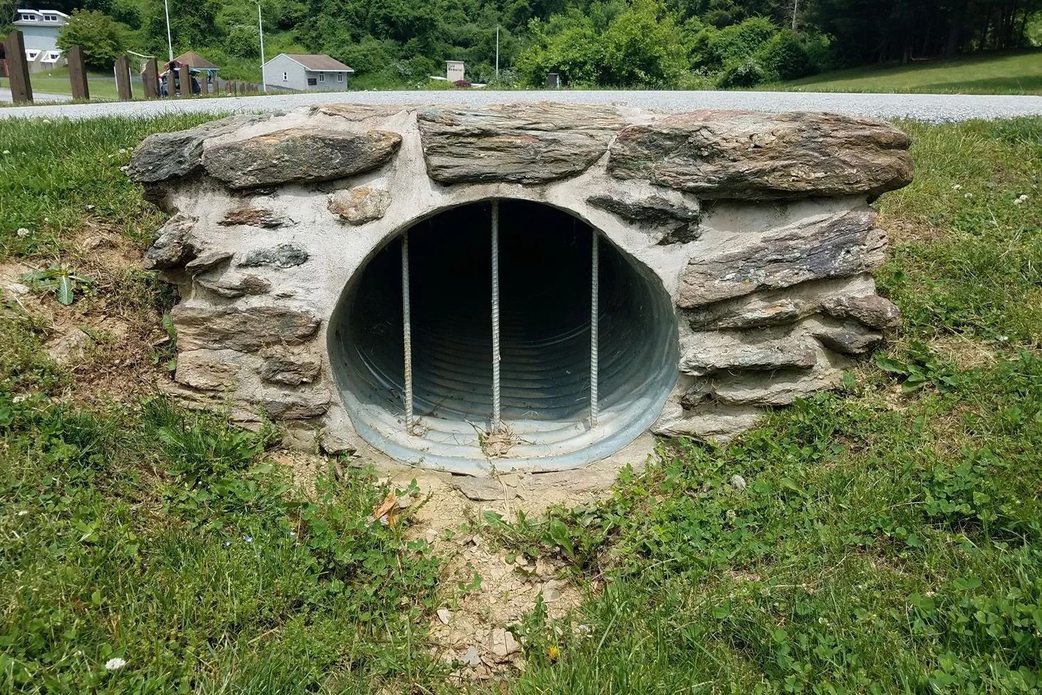A corrugated metal culvert with vertical metal bars embedded in a stone and mortar headwall set into a grassy hillside.