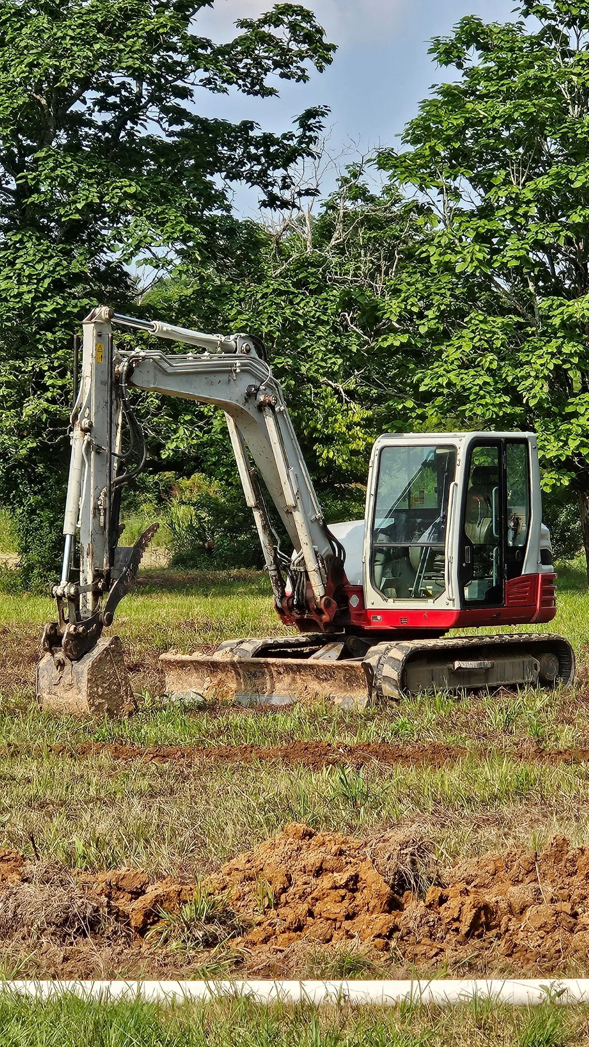A red and white Takeuchi mini excavator sits in a grassy, partially cleared field near trees.