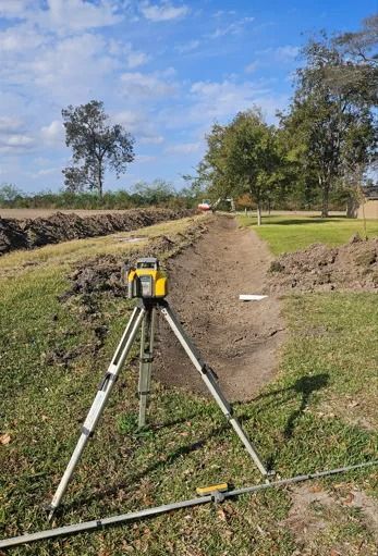 A yellow laser level on a tripod faces down a trench in a grassy, rural field under a bright, cloudy sky.