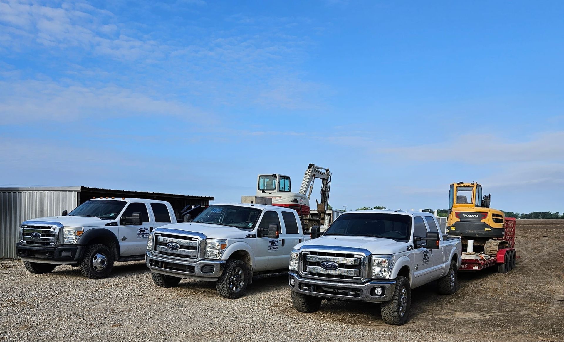 Three white Ford pickup trucks parked on a gravel lot, with a yellow excavator on a trailer nearby.