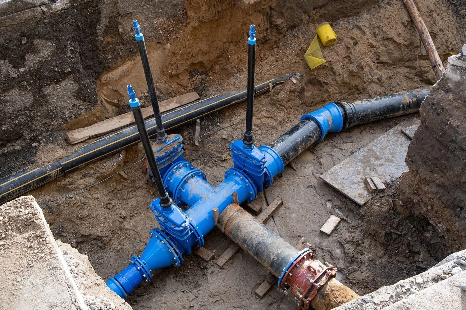A construction site showing blue pipe fittings and valves connected to dark pipes in a shallow dirt trench.