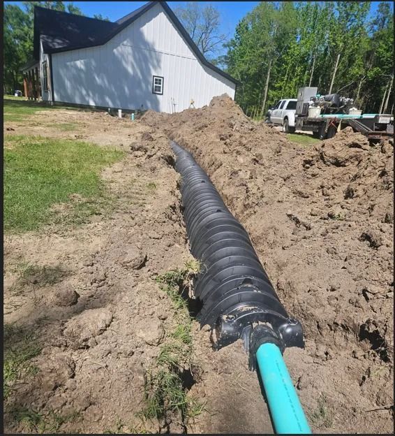 A trench in a yard containing a black corrugated drainage chamber connected to a blue PVC pipe, near a white building.