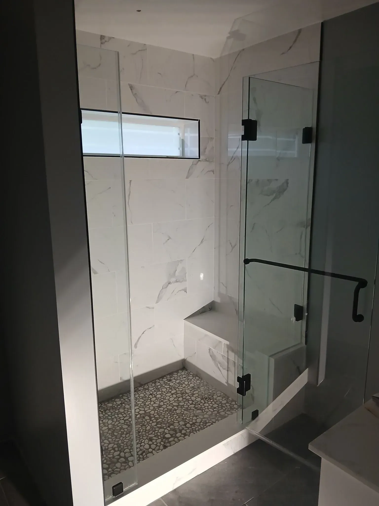 Modern walk-in shower with glass door, white marble walls, pebble floor, and a small frosted window.
