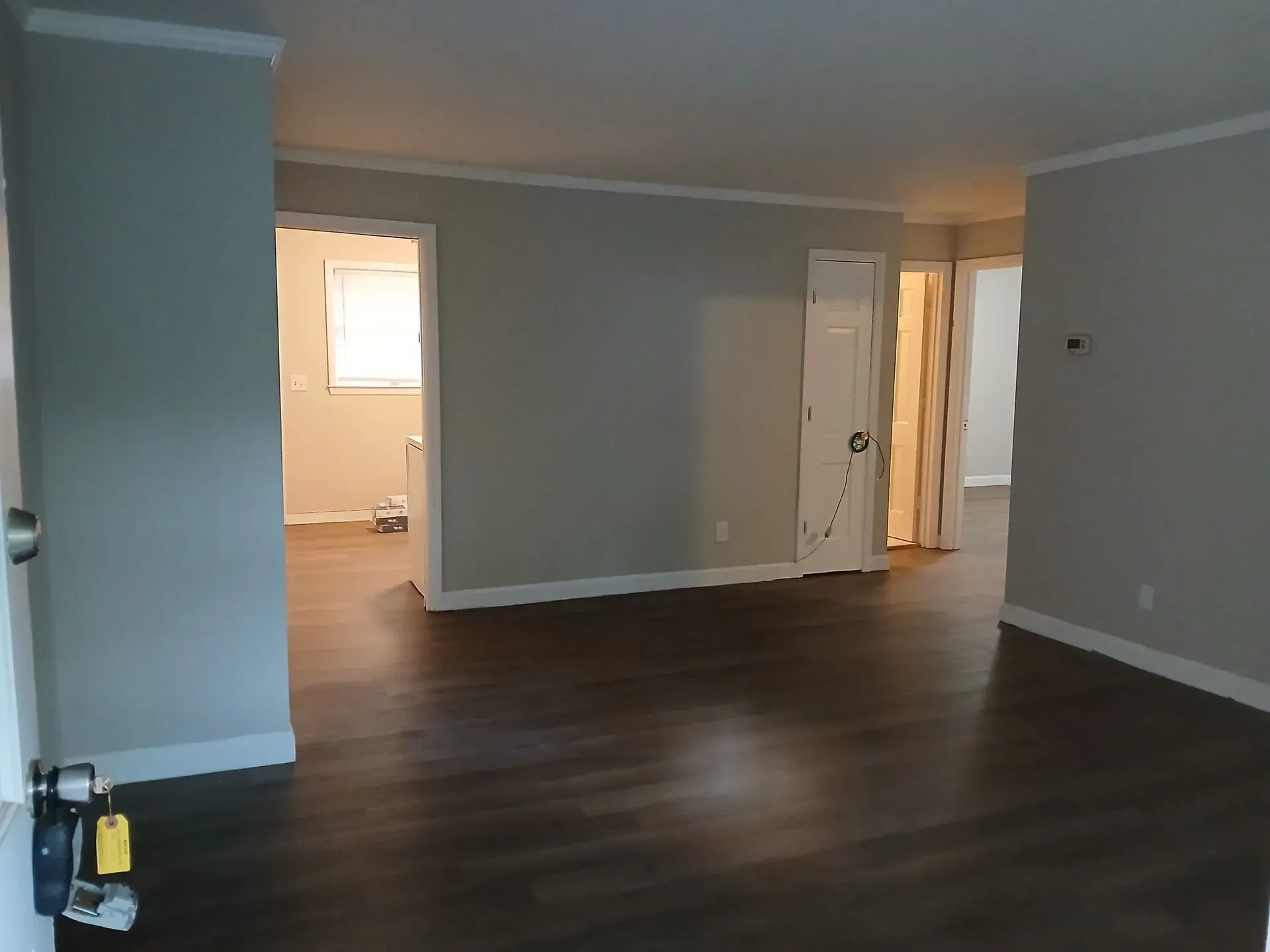Empty living room with dark wood floors, gray walls, and a doorway to a lit room beyond
