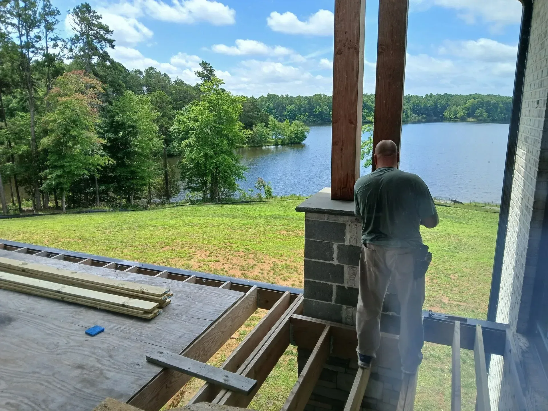 Person standing on a porch overlooking a lake and trees on a sunny day
