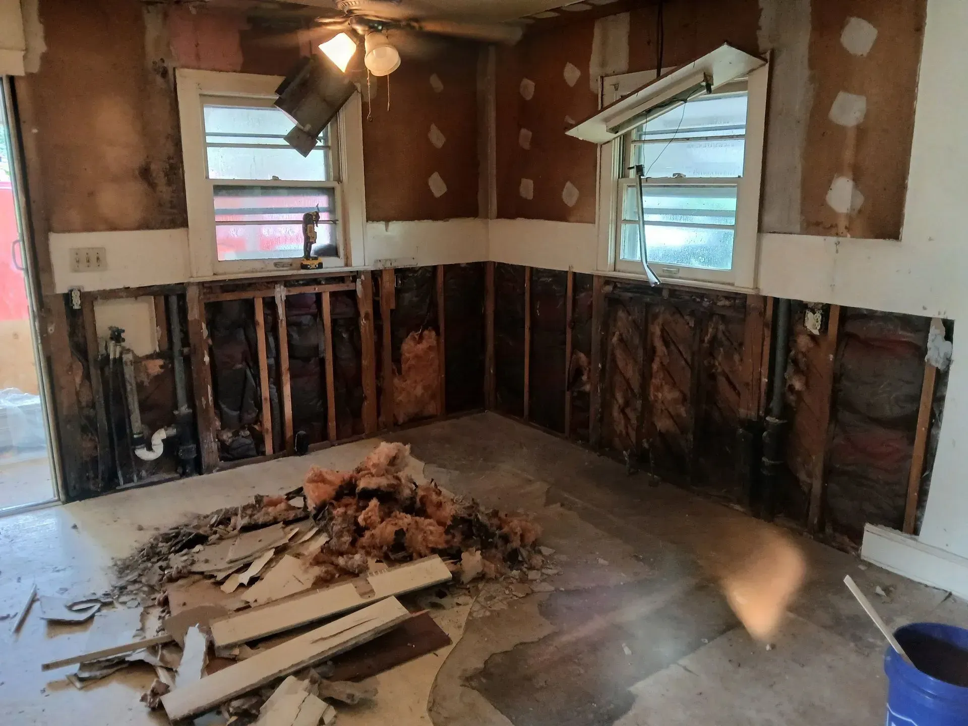 Damaged room with exposed studs, torn drywall, debris on floor, and two windows with daylight.